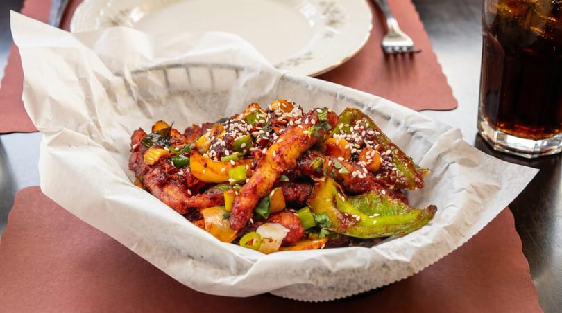 Spicy chicken with peppers and sesame seeds in a basket, with a drink and plate in the background.