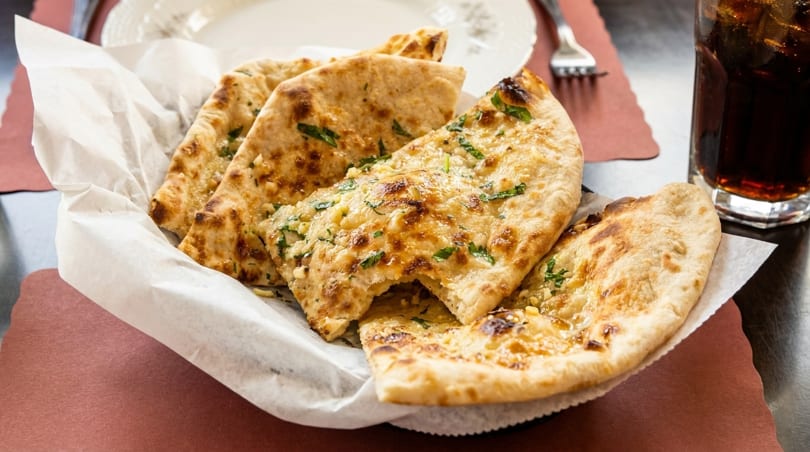 Basket of golden-brown garlic naan bread, garnished with green herbs, on a table with a drink.