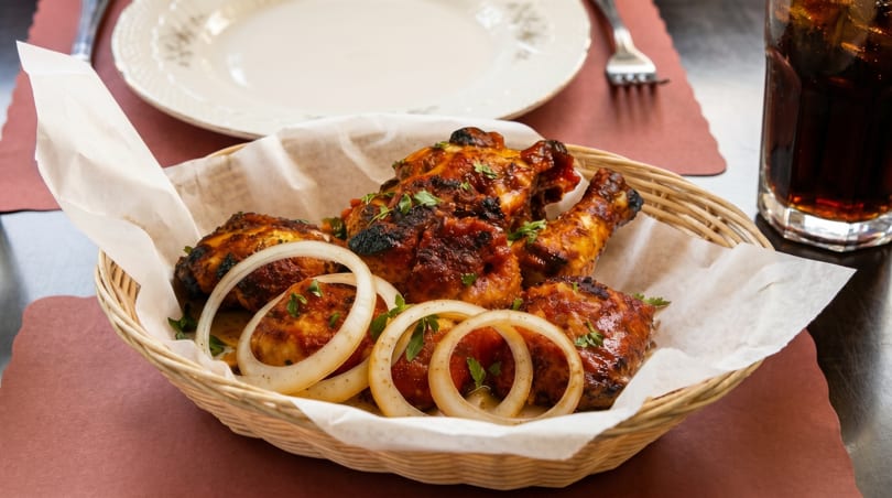 Basket of grilled chicken drumsticks and thighs, garnished with onion rings and parsley, with a drink.