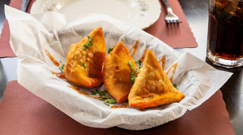 Three golden-brown samosas drizzled with sauce and garnished with cilantro in a white paper-lined basket.