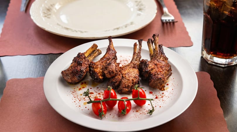 Four grilled lamb chops with cherry tomatoes on a white plate, with a drink and empty plate in the background.